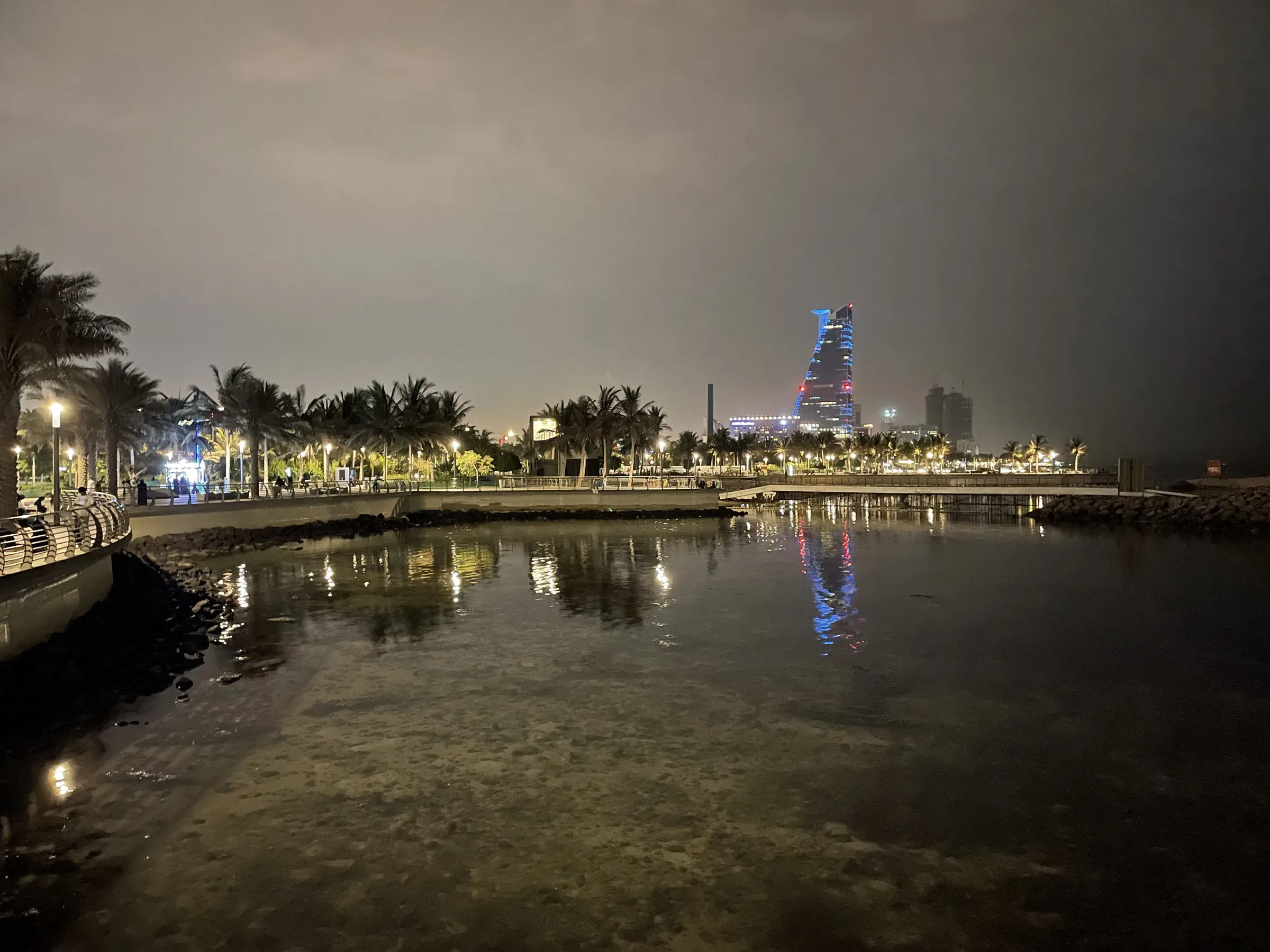 Palm-lined Jeddah Corniche promenade at night with an illuminated waterfront tower reflected in the water